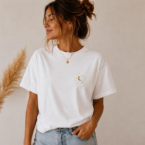 Può includere: Una t-shirt bianca a maniche corte con una piccola grafica dorata di una luna crescente circondata da minuscole stelle sul lato sinistro del petto. La maglietta ha uno scollo rotondo ed è abbinata a un paio di jeans in denim azzurro.