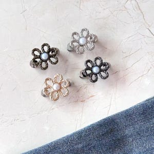 Pode incluir: Quatro botões em forma de flor com pétalas de metal torcido em tons de prata, bronze, cinza escuro e dourado. Cada botão apresenta uma conta central branca semelhante a uma pérola. Os botões estão dispostos em grupo, exibindo seu design.