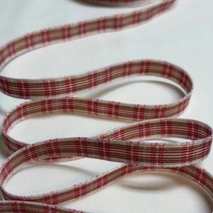 May include: A narrow fabric ribbon featuring a woven plaid pattern in shades of red, white, and tan. The ribbon is coiled in a loose, serpentine shape, displaying a consistent grid-like design across its entire length and textured surface.