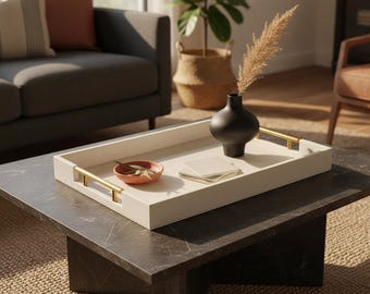 May include: A rectangular off-white serving tray with gold-toned metal handles. Inside the tray sits a matte black vase containing a dried plant stem, a small terracotta-colored bowl with green leaves, and a stack of three square white coasters.