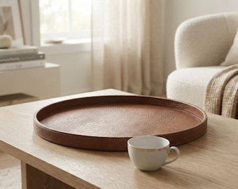 May include: A large, round, brown tray with a textured, embossed surface pattern and a raised rim, accompanied by a small, white ceramic teacup featuring a thin gold-colored rim detail, both resting on a light-toned wooden surface.
