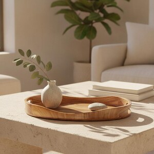 May include: An oval wooden tray with integrated handles, featuring a smooth, natural grain finish. Inside the tray sits a small, off-white ceramic bud vase containing a single green leafy stem, alongside a smooth, light-colored river stone.