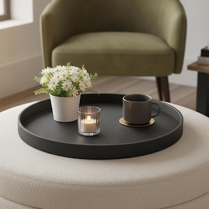 May include: A round black tray with a textured surface sits on a cream-colored ottoman. The tray holds a white pot containing small white daisy-like flowers, a clear glass votive candle holder with a lit candle, and a grey mug on a gold coaster.