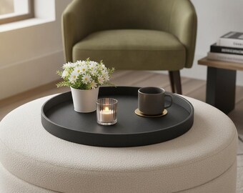May include: A round black tray with a textured surface sits on a cream-colored ottoman. The tray holds a white pot containing small white daisy-like flowers, a clear glass votive candle holder with a lit candle, and a grey mug on a gold coaster.