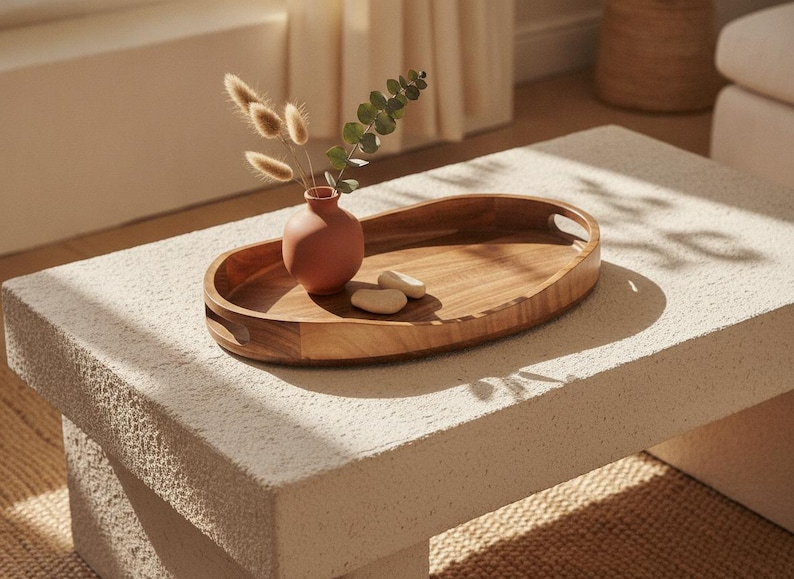 May include: An oval wooden serving tray with integrated handles, featuring a smooth, light brown grain. Inside the tray sits a small, matte terracotta-colored vase containing dried bunny tail grass and a single green eucalyptus stem.