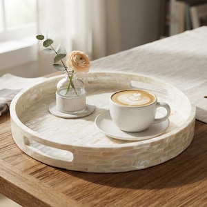 May include: A round, off-white tray with a mosaic-style shell inlay and two integrated side handles. The tray holds a small clear glass vase containing a single peach-colored flower and a white ceramic coffee cup on a matching saucer.