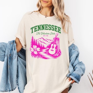 May include: A cream-colored t-shirt featuring a graphic print with the text TENNESSEE The Volunteer State EST. 1796 in green and pink. The design includes a pink illustration of mountains, pine trees, a flower, and an acoustic guitar.