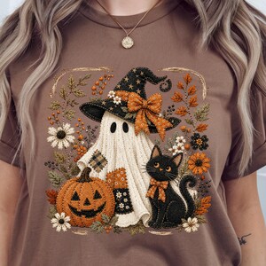 May include: A brown t-shirt featuring a graphic print of a white ghost wearing a black witch hat with an orange bow, a black cat with an orange bow, and an orange jack-o'-lantern, all surrounded by autumn-themed flowers and foliage.