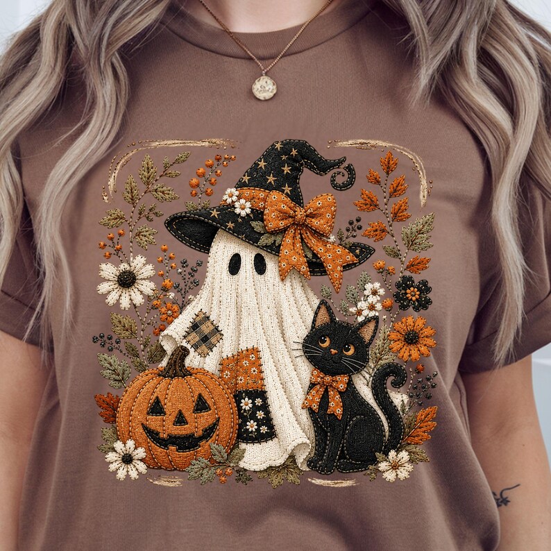 May include: A brown t-shirt featuring a graphic print of a white ghost wearing a black witch hat with an orange bow, a black cat with an orange bow, and an orange jack-o'-lantern, all surrounded by autumn-themed flowers and foliage.
