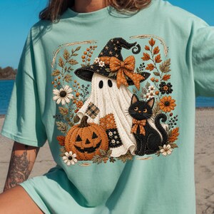 May include: A light mint green t-shirt featuring a central graphic of a white ghost wearing a black witch hat with an orange bow, a black cat with an orange bow, and an orange jack-o'-lantern, all surrounded by autumn leaves and flowers.