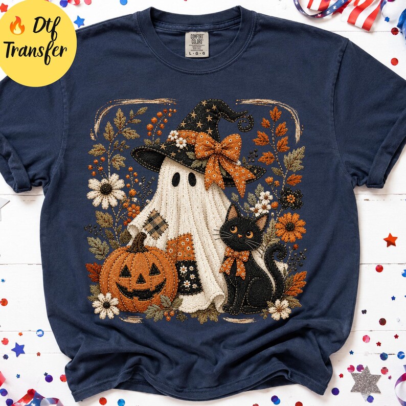 May include: A navy blue short-sleeve t-shirt featuring a graphic print of a white ghost wearing a black witch hat with an orange bow, a black cat with an orange bow, and an orange jack-o'-lantern surrounded by autumn leaves and white flowers.