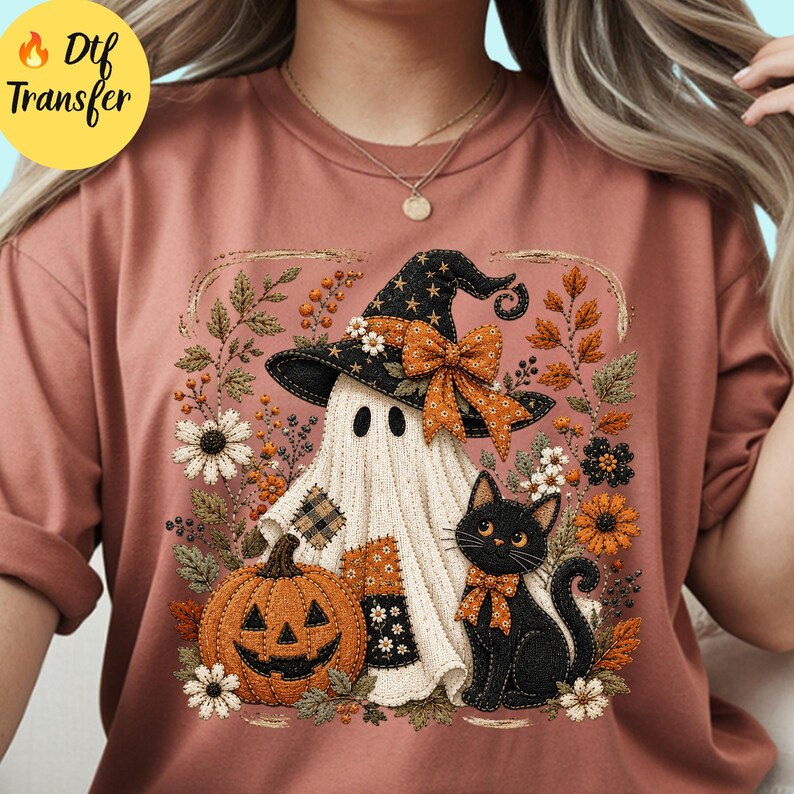 May include: A dusty rose t-shirt featuring a graphic print of a ghost wearing a black witch hat with an orange bow, a jack-o'-lantern, and a black cat with an orange bow, all surrounded by embroidered-style autumn flowers and leaves.
