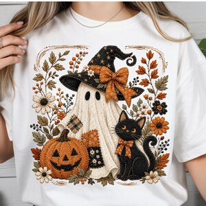 May include: A white t-shirt featuring a graphic print of a ghost wearing a black witch hat with an orange bow, a jack-o'-lantern, and a black cat with an orange bow, all surrounded by autumn-themed flowers, leaves, and berries in orange and brown.