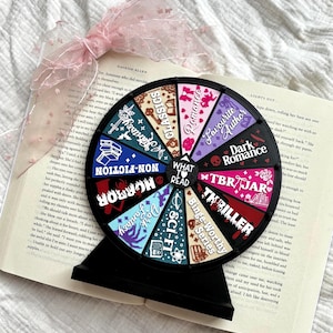 May include: A black circular decision wheel with a stand, featuring colourful segments labelled with book genres like Fantasy, Classics, Romance, Favourite Author, Dark Romance, TBR Jar, Thriller, Binge-Worthy Series, Sci-Fi, Dark Fantasy, Horror, and Non-Fiction.