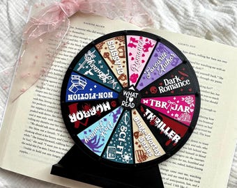 May include: A black circular decision wheel with a stand, featuring colorful segments labeled with book genres like Fantasy, Classics, Romance, Favourite Author, Dark Romance, TBR Jar, Thriller, Binge-Worthy Series, Sci-Fi, Dark Fantasy, Horror, and Non-Fiction.