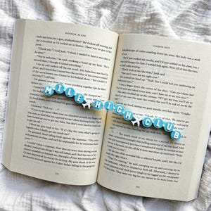 May include: A bookmark made of light blue circular beads with white letters spelling MILE HIGH CLUB, interspersed with two small white aeroplane charms, resting horizontally across the open pages of a printed paperback book.