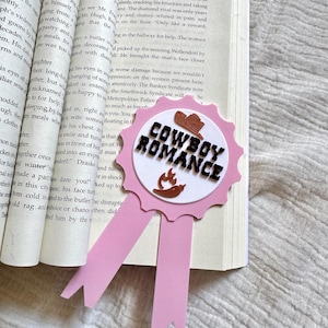 May include: A pink ribbon bookmark with a scalloped circular top featuring the text "COWBOY ROMANCE" in black letters, accompanied by a small brown cowboy hat icon and a brown chilli pepper icon with orange flames, resting on an open book page.