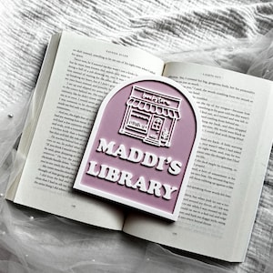 May include: A mauve-coloured, arch-shaped book marker featuring a white raised graphic of a storefront with an awning and the text "bookstore" above the words "MADDI'S LIBRARY" in bold, white, raised lettering, resting on an open book.