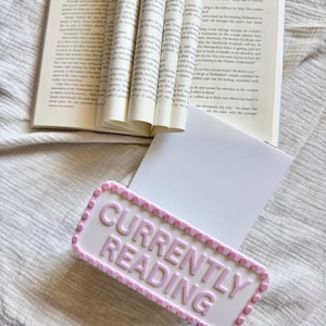 May include: A rectangular white sign with raised light pink lettering that reads CURRENTLY READING, framed by a border of small pink circles. The sign is positioned in front of an open book with visible printed text on the pages.