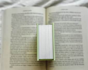 May include: A small, rectangular object with a light green frame and a series of vertical white accordion-folded paper panels, positioned upright in the centre of an open book with visible printed text on the pages.