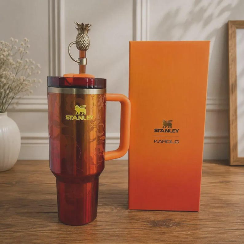 May include: A metallic orange and red gradient Stanley tumbler with a matching orange handle and lid. A gold-toned pineapple topper sits on the straw. The tumbler features a floral pattern and the Stanley logo. Beside it stands an orange box with Stanley and KAROL G branding.