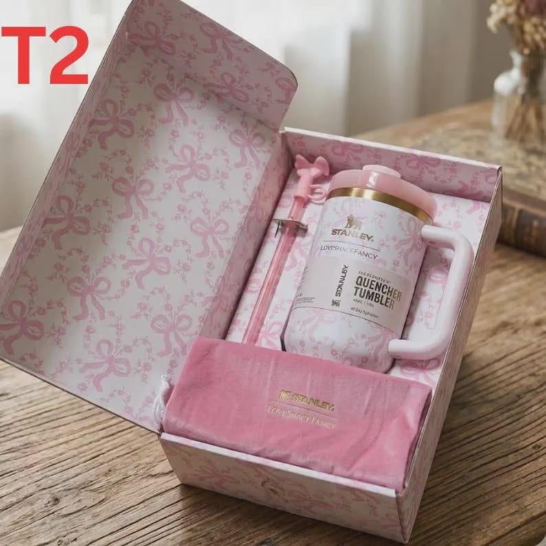 May include: A pink and white gift box featuring a bow pattern containing a Stanley Quencher tumbler with a matching pink straw and a pink velvet pouch. The tumbler is labeled with the text Stanley, LoveShackFancy, and Quencher Tumbler.