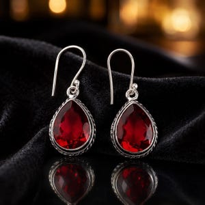 Pode incluir: Um par de brincos pendentes com pedras vermelho-escuras facetadas em forma de pera, engastadas em molduras de prata com uma borda de corda torcida. Os brincos pendem de ganchos de prata simples e exibem um acabamento polido nas pedras.