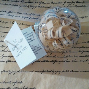 May include: A clear glass ornament filled with rolled up pages of the US Constitution. The ornament is sitting on a piece of parchment paper with the text "The Constitution of the United States" printed on a white tag.