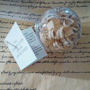 May include: A clear glass ornament filled with curled pages of the US Constitution. The ornament is labeled "The Constitution of the United States".
