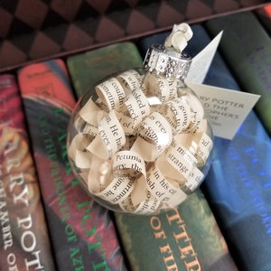 May include: A clear glass Christmas ornament filled with curled paper strips, resting on a collection of Harry Potter books. The ornament has a silver cap and is filled with text from the book. The books are in various colors.