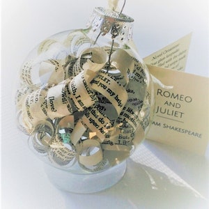 Peut inclure: Une boule de No&euml;l en verre transparent remplie de pages roul&eacute;es de la pi&egrave;ce Rom&eacute;o et Juliette de William Shakespeare. La boule a un sommet argent&eacute; et repose sur une surface blanche.