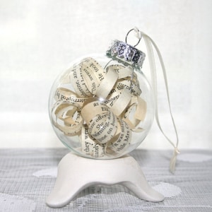 Könnte beinhalten: Ein transparenter Glas-Weihnachtsbaumschmuck, gefüllt mit aufgerollten Buchseiten. Der Schmuck steht auf einem weißen Ständer und hat eine silberne Spitze mit einem weißen Band.