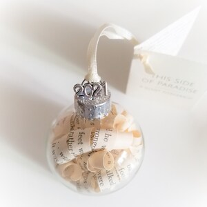 May include: A clear glass ornament filled with rolled up pieces of paper. The ornament has a silver top with the year 2021 on it. The ornament is hanging from a white ribbon. A white tag with the text "This Side of Paradise" is attached to the ribbon.