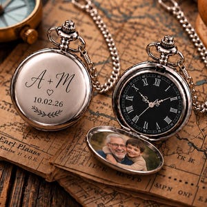 Peut inclure: Deux montres de poche argentées avec chaînes. L'une est fermée, gravée avec le texte A + M, un cœur, la date 10.02.26 et un motif de feuilles. L'autre est ouverte, montrant un cadran noir avec des chiffres romains et une photo de deux personnes.