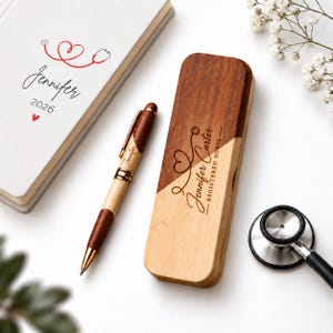 Puede incluir: Un bolígrafo de madera y un estuche rectangular de madera a juego, ambos grabados con el nombre Jennifer Carter y un diseño de corazón con estetoscopio. El bolígrafo tiene un acabado de madera en dos tonos con detalles dorados y el estuche dice Registered Nurse.