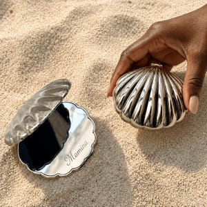 May include: A silver-toned, clam-shaped compact mirror with a hinged lid. One half of the shell is closed, while the other half is open to reveal a reflective mirror surface with the name Mamina engraved in a cursive script on the lower edge.