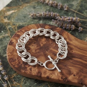 May include: A silver chain link bracelet featuring a toggle clasp closure. The bracelet is constructed from a series of interlocking circular metal rings, creating a continuous circular band with a polished, reflective metallic finish.
