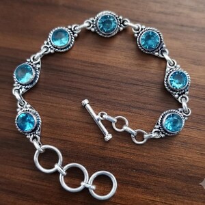 Op de afbeelding: Een zilveren armband met zeven ronde, gefacetteerde blauwe stenen in decoratieve metalen zettingen. De schakels zijn verbonden door kleine zilveren ringen en het sieraad wordt gesloten met een staaf- en ringvormige knevelsluiting.