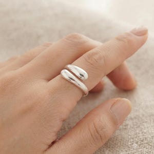 Könnte beinhalten: Ein polierter silberfarbener Ring im Bypass-Design mit zwei glatten, abgerundeten, tropfenförmigen Enden, die sich leicht überlappen. Das Metall hat eine reflektierende Hochglanzoberfläche und eine durchgehende, fließende, offene Ringstruktur.