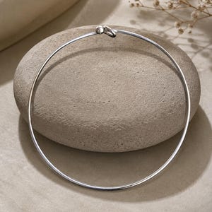 May include: A thin, circular silver-toned metal necklace featuring a simple hook-and-eye clasp mechanism. The necklace is formed from a single continuous wire that maintains a rigid, rounded shape, showcasing a polished, reflective metallic surface.