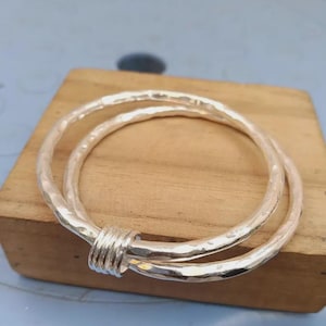 Op de afbeelding: Een zilveren armband bestaande uit twee in elkaar grijpende cirkelvormige banden met een gehamerde, getextureerde afwerking. De twee banden zijn verbonden door een kleine, gladde zilveren verbindingsring die ze in een vaste positie houdt.