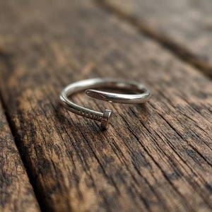 May include: A silver-toned metal ring shaped like a bent nail. The ring features a smooth, polished finish with a rounded head at one end and a tapered point at the other. The design is open-ended, allowing for adjustment, and has a metallic sheen.