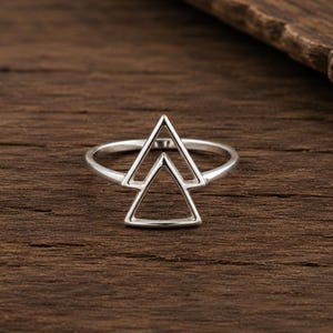 Peut inclure: Une bague en argent poli présentant un motif géométrique composé de deux triangles ouverts superposés. L'anneau est fin et circulaire, soutenant la pièce centrale triangulaire symétrique qui se dresse verticalement.