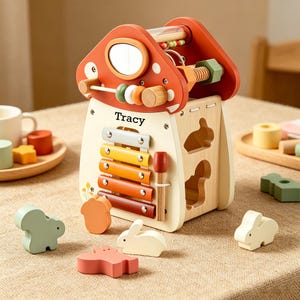 May include: A wooden activity cube shaped like a mushroom with the name Tracy printed on the front. It features a xylophone, a mirror, a bead maze, and shape-sorting cutouts. Several wooden animal-shaped blocks are scattered in front of the toy.