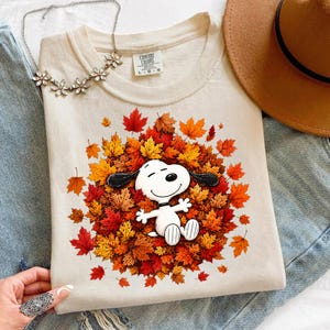 May include: A cream-colored t-shirt featuring a graphic of the cartoon character Snoopy lying in a pile of vibrant autumn leaves in shades of red, orange, and yellow. The shirt has a crew neckline and a Comfort Colors brand label at the inner collar.