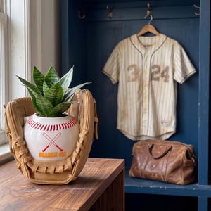 Peut inclure: Un gant de baseball en cuir beige tenant un pot en céramique blanc en forme de balle de baseball avec des coutures rouges, un motif rouge de deux battes croisées et le texte "GRAHAM 610" en orange, contenant une plante serpent verte.