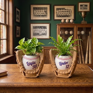 Peut inclure: Deux pots de fleurs en céramique en forme de balle de baseball nichés dans des gants de baseball en cuir beige. L'un porte le nom Layla et le numéro 98 en bleu, l'autre le nom Lindor et le numéro 12 en bleu.
