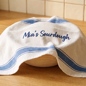 May include: A white kitchen towel with blue stripes draped over a round proofing basket. The towel features the embroidered text "Mia's Sourdough" in blue script lettering. The fabric has a textured weave and hangs down over the sides of the basket.