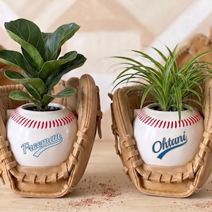 Peut inclure: Deux pots de fleurs en céramique en forme de balles de baseball avec des coutures rouges, chacun niché dans un gant de baseball en cuir beige. Un pot porte le nom Freeman en bleu, l'autre le nom Ohtani en bleu, contenant chacun une plante verte.