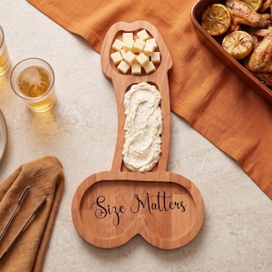 May include: A light brown wooden serving board shaped like a phallus with three distinct sections. The bottom section features the engraved text "Size Matters" in a cursive font. The top two sections contain cubed cheese and a creamy white dip.